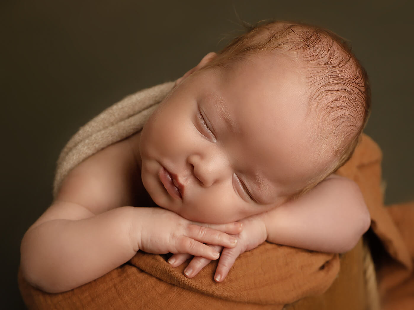 Sleeping newborn baby resting its head on folded arms, wrapped in a warm brown blanket.